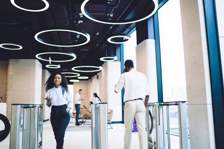 Content coworkers going through access control at workplace Low angle of glad African American staff in stylish clothes passing through turnstile while using smartphone in hall of contemporary business center