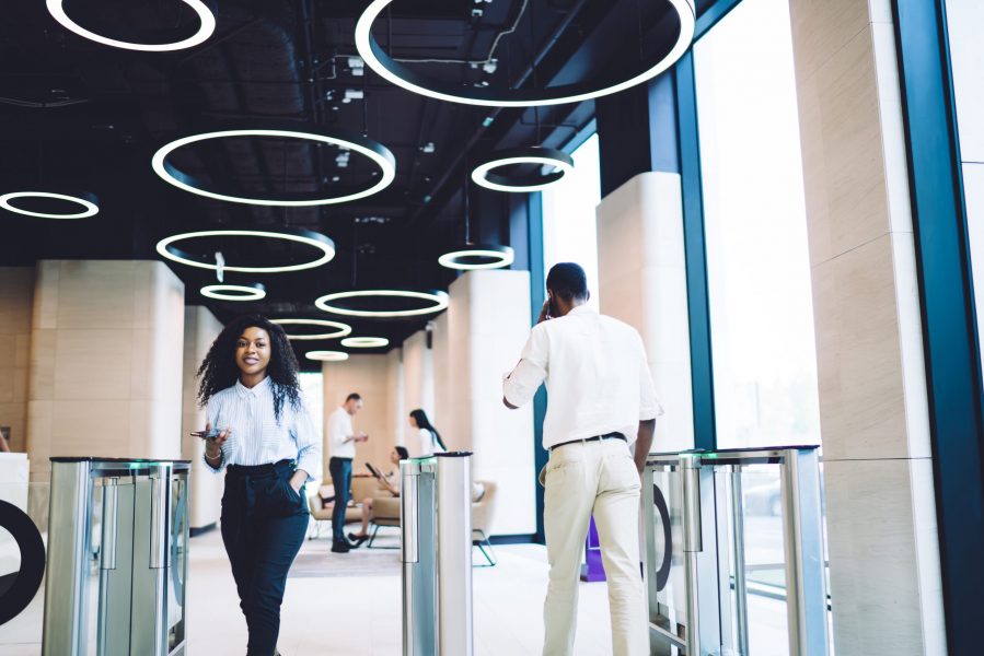 Low angle of glad African American staff in stylish clothes passing through turnstile while using smartphone in hall of contemporary business center